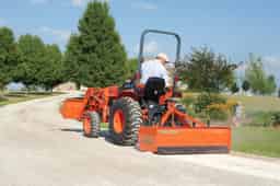 Tractor-mounted 60-inch box blade attachment leveling a gravel driveway with operator seated on orange compact utility tractor on a sunny rural property