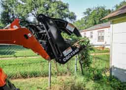 Orange skid-steer with a 40" black McCullough grapple bucket (model CGC1040) picking up tree branches and yard debris in a residential backyard near a chain-link fence and houses