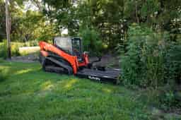 Compact orange Kubota track loader fitted with a 72-inch black brush mower attachment cutting dense roadside brush and tall grass beside trees and a gravel driveway