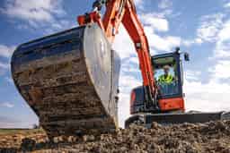 Excavator operator in high-visibility vest using a large dirt-filled bucket attachment on an orange excavator at a construction site