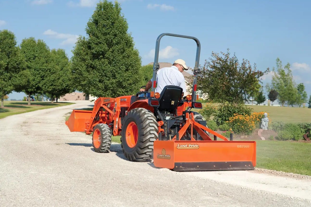 Tractor-mounted 60-inch box blade attachment leveling a gravel driveway with operator seated on orange compact utility tractor on a sunny rural property
