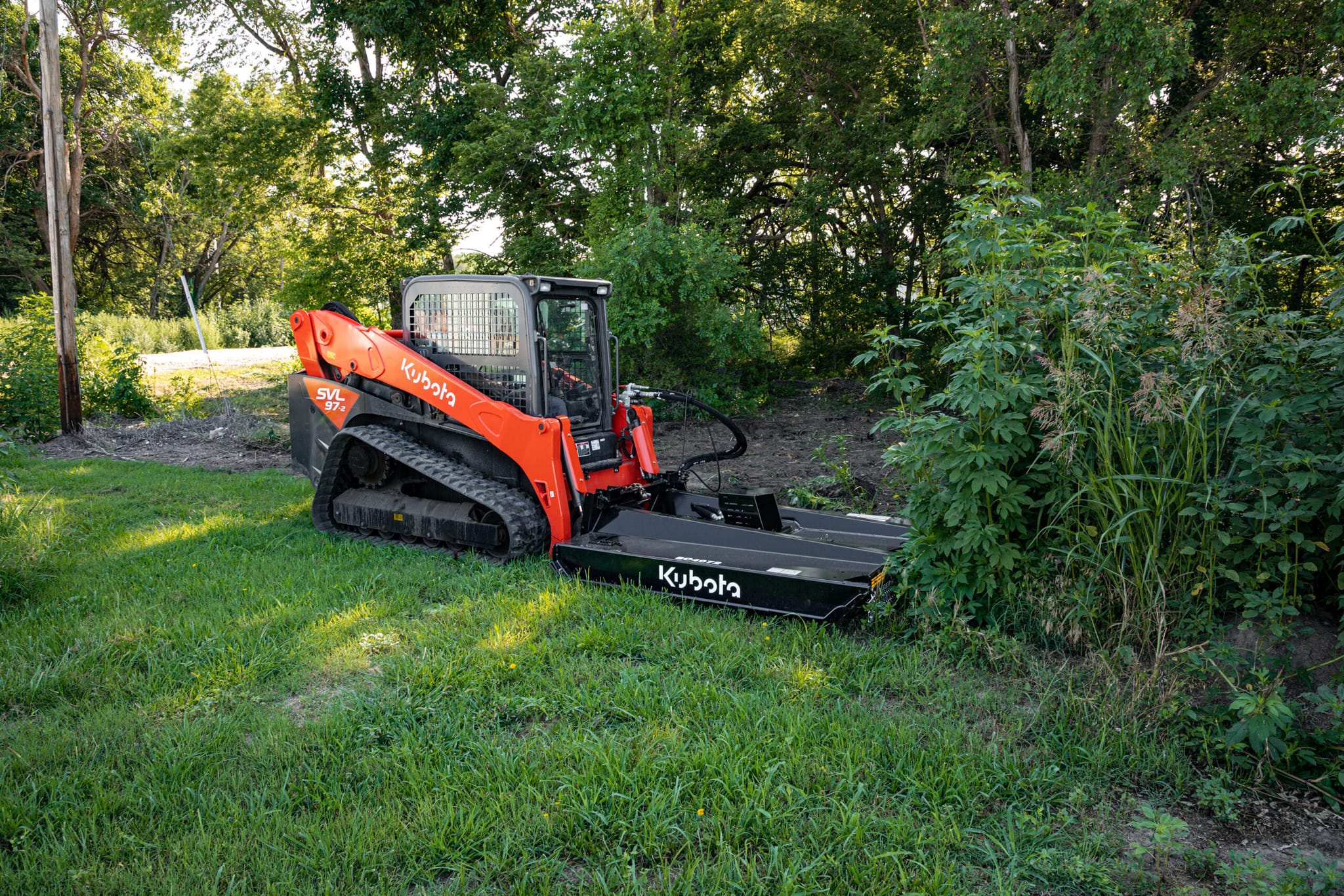 Compact orange Kubota track loader fitted with a 72-inch black brush mower attachment cutting dense roadside brush and tall grass beside trees and a gravel driveway