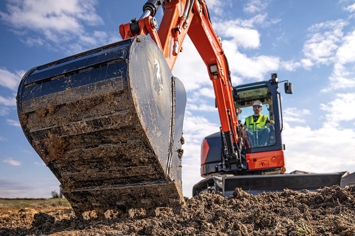 Excavator operator in high-visibility vest using a large dirt-filled bucket attachment on an orange excavator at a construction site